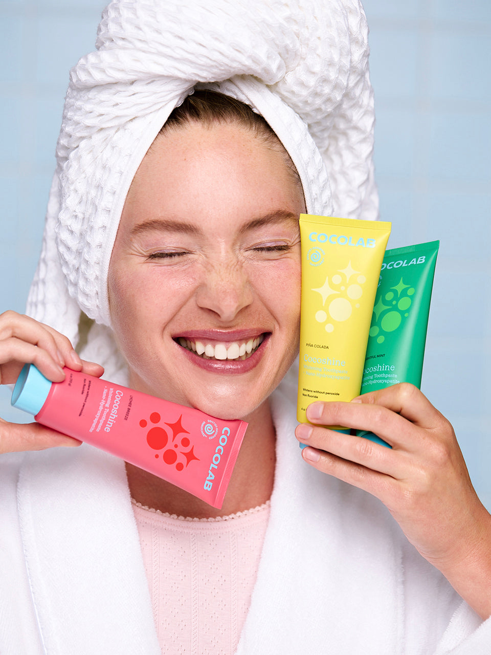 Woman with a towel on her head holding three Cocoshine toothpaste tubes against a light blue background