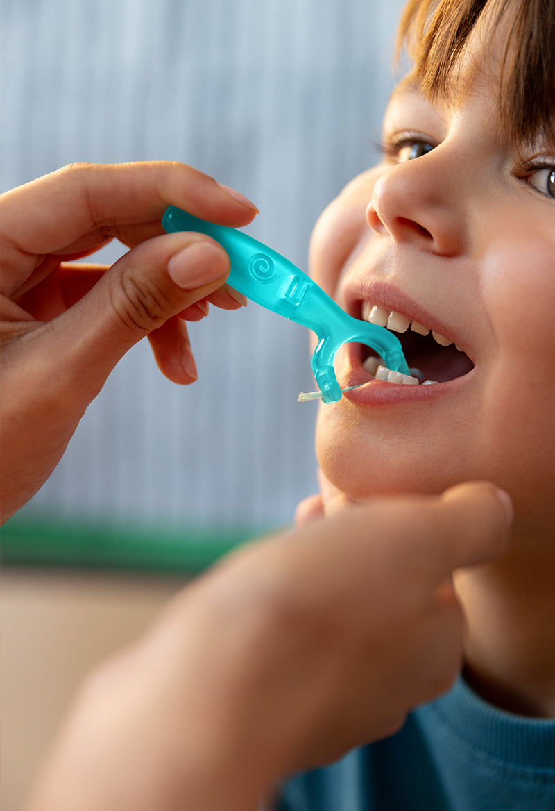 Child being assisted with a blue toothbrush by an adult hand against a neutral background