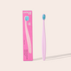 Pink toothbrush with blue bristles next to its packaging on a light pink background